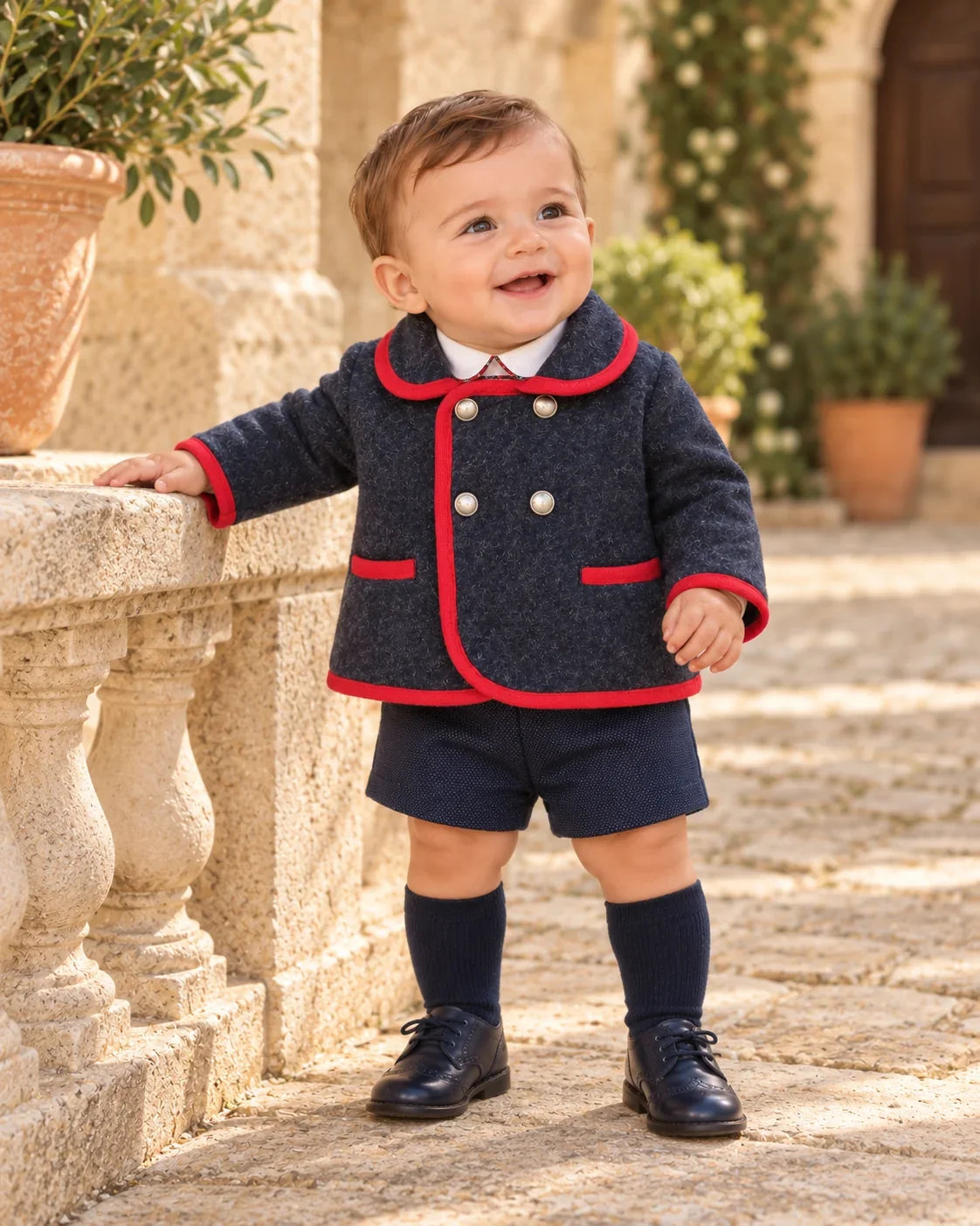 Baby boy wearing a navy coat with red trim, navy shorts and knee-high socks for autumn