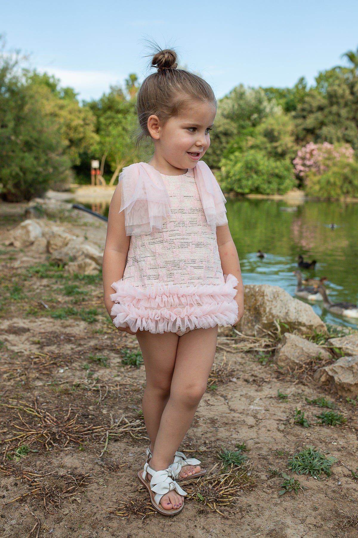 Young girl in a pink dress standing near a pond with trees in the background