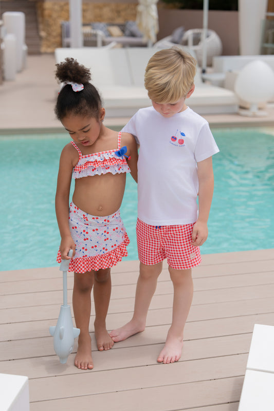 Boy wearing the Miranda Mon Petit Cherry Boys T-Shirt and red gingham swimming trunks beside a girl by the swimming pool.
