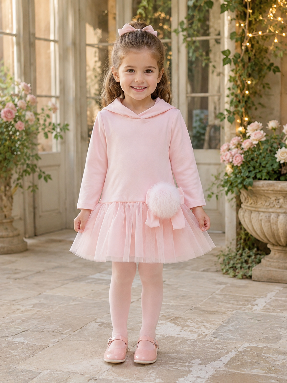 Little girl wearing Bimbalò girls pink hooded dress with tulle skirt, satin bow and faux fur pom pom detail.