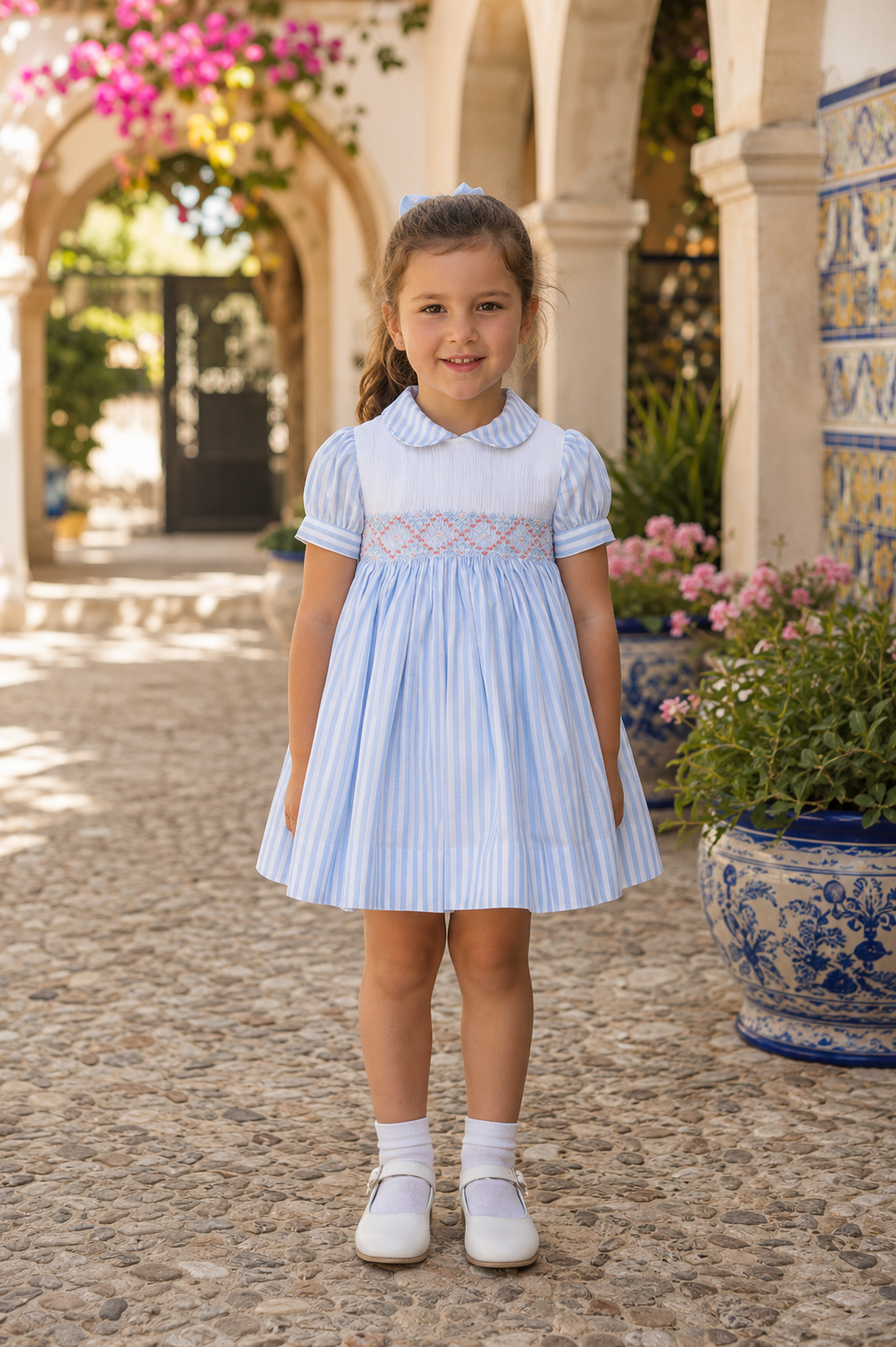Little girl wearing Naxos Carmen girls blue and white stripe cotton dress with pink hand smocking and back bow, handmade in Spain.