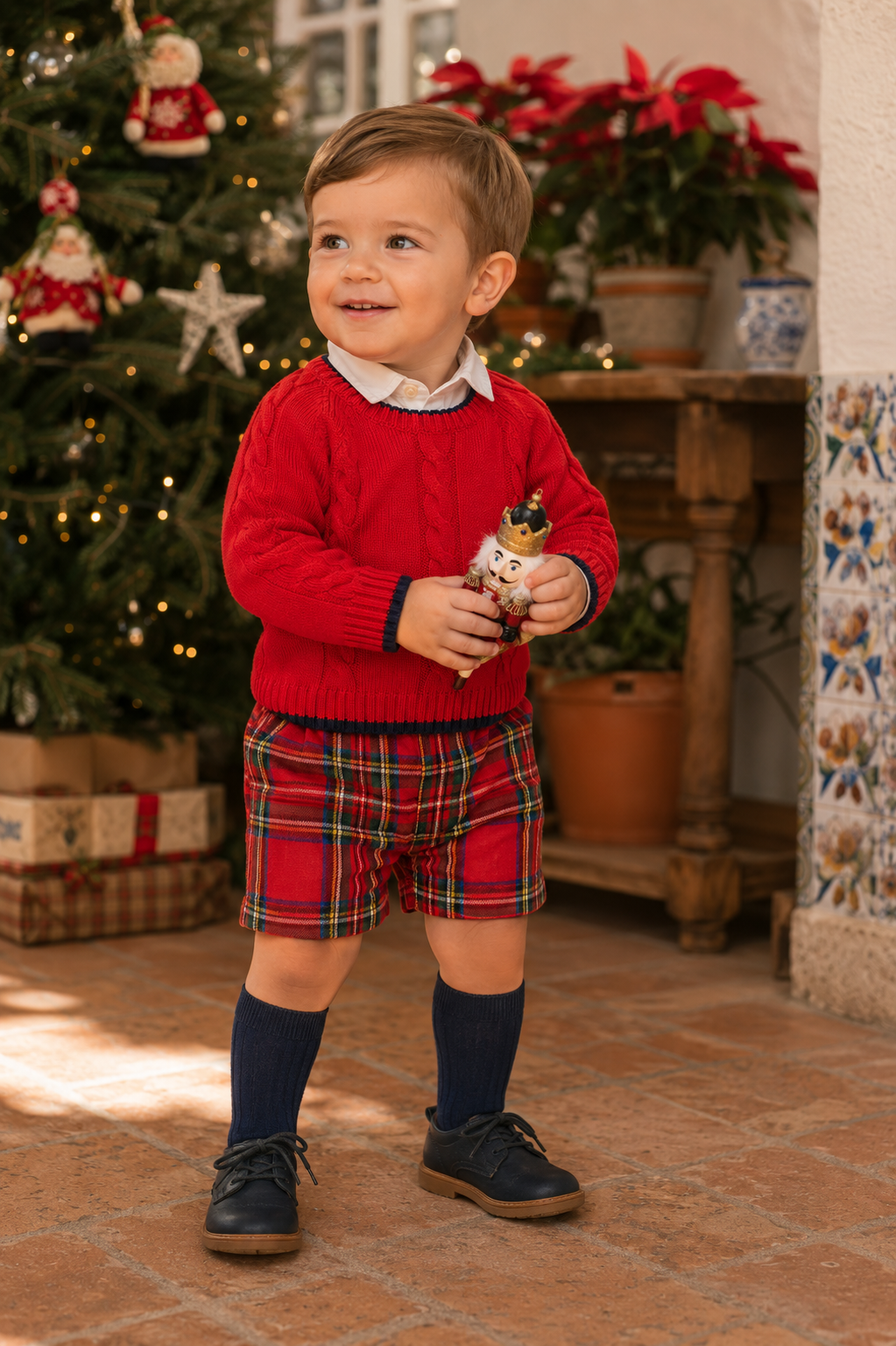 Toddler boy wearing Tutto Piccolo red tartan short set with red cable knit jumper in festive Spain setting