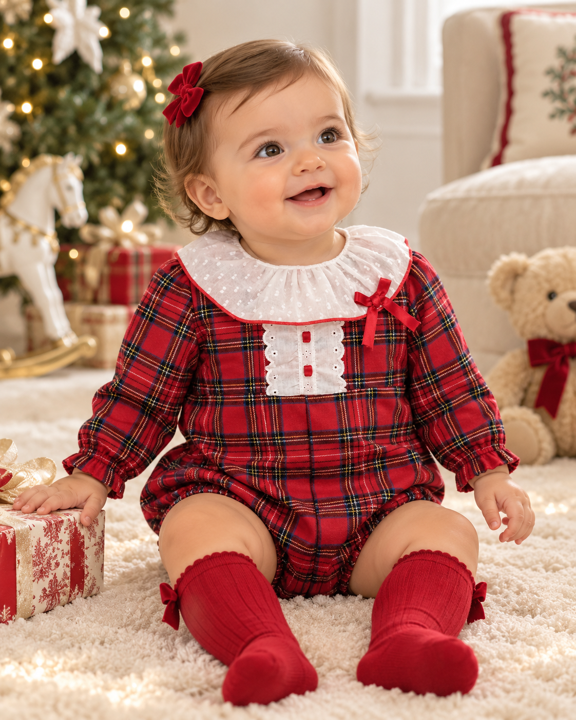 Eva Class Natividad girls red tartan romper with white dotted collar, red bow detail and long sleeves, shown on a baby girl in a festive Christmas setting