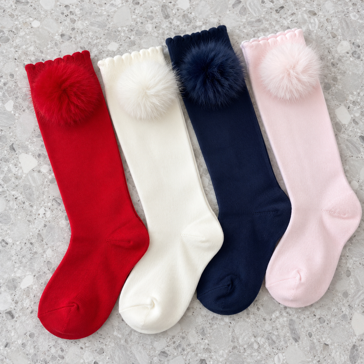 Dolce Petit girls knee-high Minky pom pom socks in red, cream, navy and pale pink, shown in a flat-lay on a grey terrazzo background