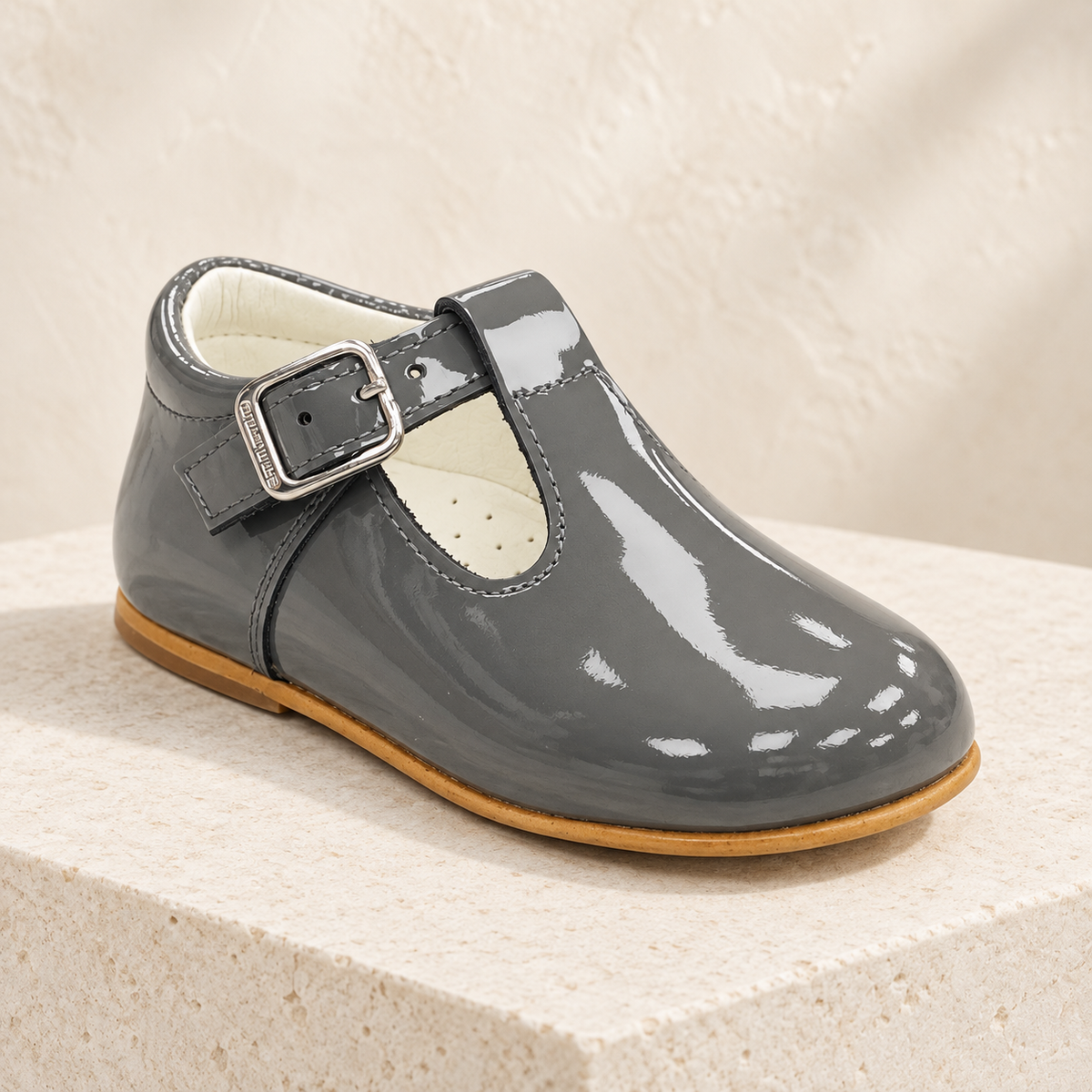 Andanines grey patent leather boys T-bar shoe on a cream stone plinth