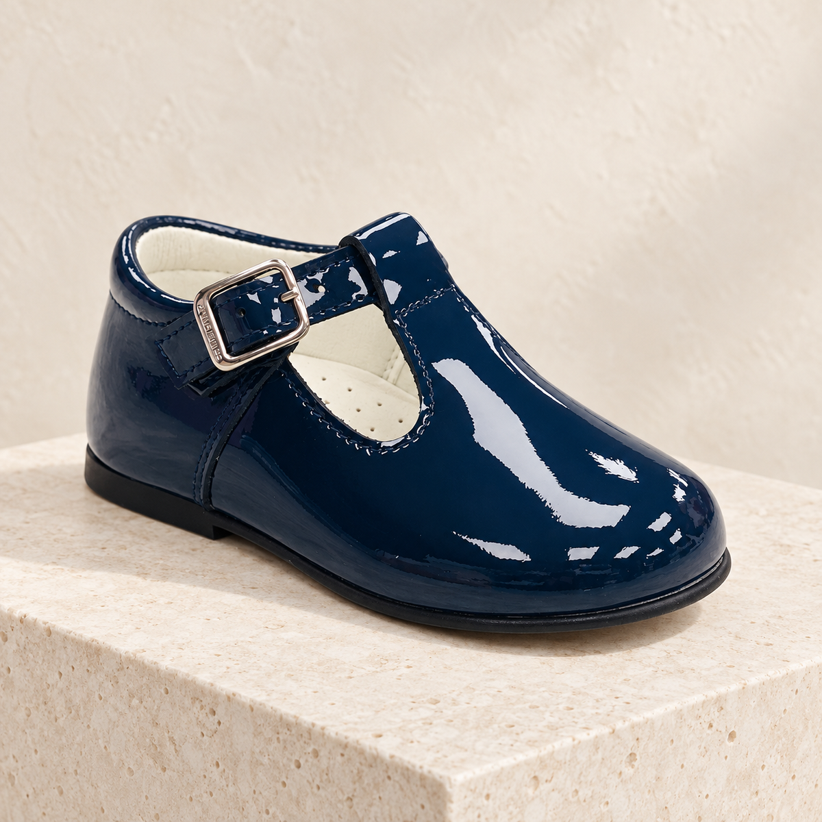 Andanines navy patent leather boys T-bar shoe on a cream stone plinth