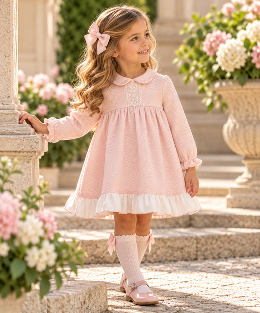 Girl modelling LOR Miral Elisa blush pink textured jacquard dress with ivory embroidered lace panel, Peter Pan collar and contrasting cream ruffle hem in a Spanish-style garden courtyard